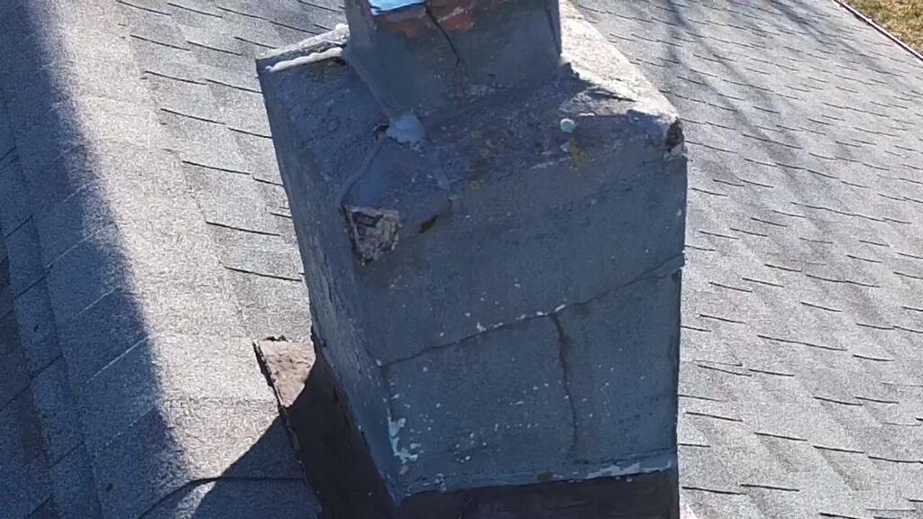 Weathered gray chimney with a cracked surface and peeling paint beside the roof