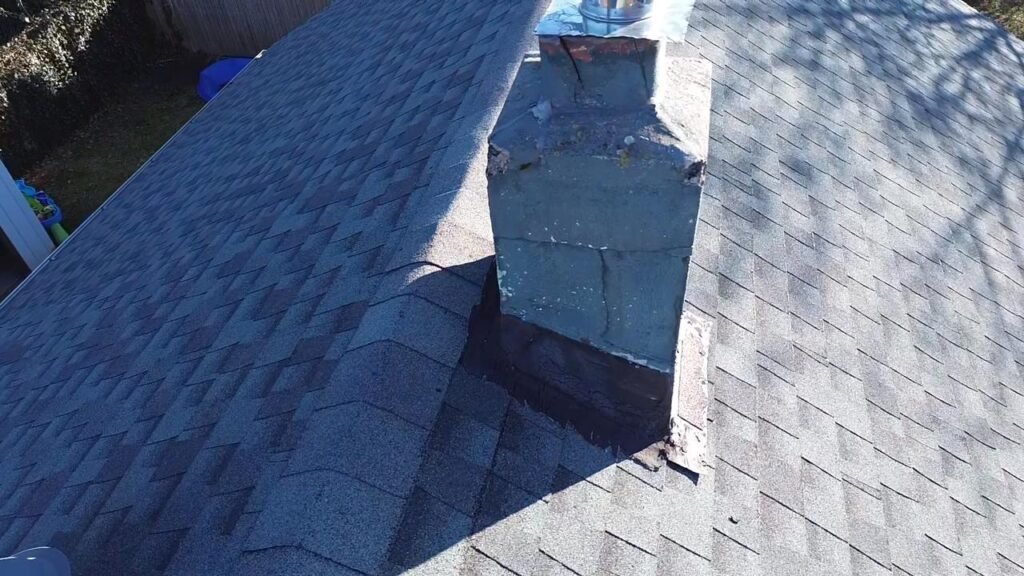 Weathered gray chimney with cracked flashing, seen from across the roof